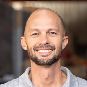 A bald man smiling in front of a warehouse.